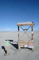A minimalist electric vehicle charging station is situated in a barren, sandy landscape with a bright blue sky overhead. The charging station is marked by various warning signs, including 'High Voltage' and 'Danger Keep Out.' Nearby, a small sign indicates it's an electric vehicle charging point.
