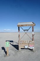 A minimalist electric vehicle charging station is situated in a barren, sandy landscape with a bright blue sky overhead. The charging station is marked by various warning signs, including 'High Voltage' and 'Danger Keep Out.' Nearby, a small sign indicates it's an electric vehicle charging point.