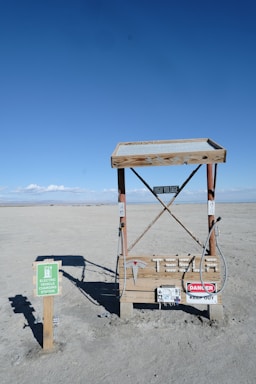 A minimalist electric vehicle charging station is situated in a barren, sandy landscape with a bright blue sky overhead. The charging station is marked by various warning signs, including 'High Voltage' and 'Danger Keep Out.' Nearby, a small sign indicates it's an electric vehicle charging point.