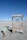 A minimalist electric vehicle charging station is situated in a barren, sandy landscape with a bright blue sky overhead. The charging station is marked by various warning signs, including 'High Voltage' and 'Danger Keep Out.' Nearby, a small sign indicates it's an electric vehicle charging point.