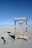 A minimalist electric vehicle charging station is situated in a barren, sandy landscape with a bright blue sky overhead. The charging station is marked by various warning signs, including 'High Voltage' and 'Danger Keep Out.' Nearby, a small sign indicates it's an electric vehicle charging point.
