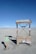A minimalist electric vehicle charging station is situated in a barren, sandy landscape with a bright blue sky overhead. The charging station is marked by various warning signs, including 'High Voltage' and 'Danger Keep Out.' Nearby, a small sign indicates it's an electric vehicle charging point.
