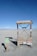 A minimalist electric vehicle charging station is situated in a barren, sandy landscape with a bright blue sky overhead. The charging station is marked by various warning signs, including 'High Voltage' and 'Danger Keep Out.' Nearby, a small sign indicates it's an electric vehicle charging point.