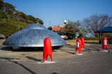 A large, metallic, saucer-shaped structure is placed on a paved area, resembling a classic UFO. Surrounding the structure are tall, red, rocket-like objects with grey circular details. Trees and greenery are in the background, along with a building featuring a red roof.