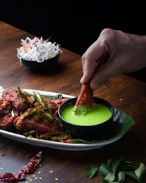 A hand dipping a piece of red, spicy fried chicken into a small black bowl of vibrant green sauce. The chicken is garnished with sliced green chilies and bits of spice. The dish is served on a white plate lined with a banana leaf, on a dark wooden table. In the background, a small bowl contains a mixed salad of shredded white and purple cabbage with thin strips of carrot.