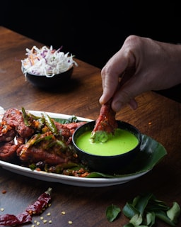 A hand dipping a piece of red, spicy fried chicken into a small black bowl of vibrant green sauce. The chicken is garnished with sliced green chilies and bits of spice. The dish is served on a white plate lined with a banana leaf, on a dark wooden table. In the background, a small bowl contains a mixed salad of shredded white and purple cabbage with thin strips of carrot.