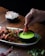 A hand dipping a piece of red, spicy fried chicken into a small black bowl of vibrant green sauce. The chicken is garnished with sliced green chilies and bits of spice. The dish is served on a white plate lined with a banana leaf, on a dark wooden table. In the background, a small bowl contains a mixed salad of shredded white and purple cabbage with thin strips of carrot.