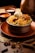 A dish of biryani topped with a boiled egg and pieces of chicken is placed in a hammered metal bowl on an orange plate. The setup is accompanied by two small bowls containing sauces, likely curry and yogurt based. Surrounding the dish are various whole spices like star anise, cardamom, and bay leaves, arranged elegantly on a wooden surface, enhancing the rustic and aromatic setting.