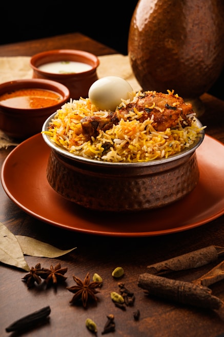 A dish of biryani topped with a boiled egg and pieces of chicken is placed in a hammered metal bowl on an orange plate. The setup is accompanied by two small bowls containing sauces, likely curry and yogurt based. Surrounding the dish are various whole spices like star anise, cardamom, and bay leaves, arranged elegantly on a wooden surface, enhancing the rustic and aromatic setting.