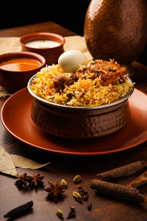 A dish of biryani topped with a boiled egg and pieces of chicken is placed in a hammered metal bowl on an orange plate. The setup is accompanied by two small bowls containing sauces, likely curry and yogurt based. Surrounding the dish are various whole spices like star anise, cardamom, and bay leaves, arranged elegantly on a wooden surface, enhancing the rustic and aromatic setting.
