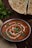 A bowl of rich, creamy paneer curry garnished with coriander leaves and a swirl of cream sits on a table. Beside it, there is a naan bread, partially sliced and garnished with herbs. Fresh cilantro leaves are placed on the table, adding a touch of green to the scene.