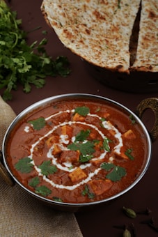 A bowl of rich, creamy paneer curry garnished with coriander leaves and a swirl of cream sits on a table. Beside it, there is a naan bread, partially sliced and garnished with herbs. Fresh cilantro leaves are placed on the table, adding a touch of green to the scene.