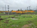 A railway yard with multiple tracks, overhead power lines, and yellow maintenance vehicles. The ground is green with vegetation and puddles, suggesting recent rain. There are utility poles and some buildings in the background.