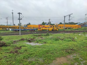 A railway yard with multiple tracks, overhead power lines, and yellow maintenance vehicles. The ground is green with vegetation and puddles, suggesting recent rain. There are utility poles and some buildings in the background.