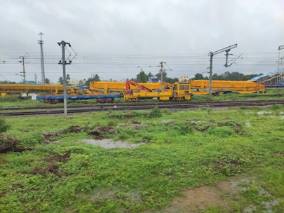 A railway yard with multiple tracks, overhead power lines, and yellow maintenance vehicles. The ground is green with vegetation and puddles, suggesting recent rain. There are utility poles and some buildings in the background.