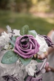 A beautifully arranged bouquet of artificial flowers, featuring prominently a purple rose adorned with two rings on top. The bouquet also includes white flowers and green leaves, set against a softly blurred natural background. The overall composition suggests a romantic or ceremonial theme.