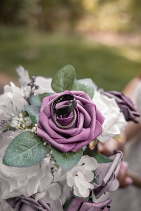 A beautifully arranged bouquet of artificial flowers, featuring prominently a purple rose adorned with two rings on top. The bouquet also includes white flowers and green leaves, set against a softly blurred natural background. The overall composition suggests a romantic or ceremonial theme.