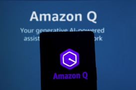 A silhouetted smartphone displays the Amazon Q logo against a blurred blue background with text. The logo is hexagonal with a stylized 'Q' in purple. The background text refers to a generative AI-powered assistant.