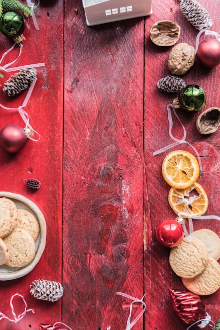 A rustic red wooden table serves as a festive backdrop, adorned with pinecones, walnuts, dried orange slices, and Christmas ornaments in shades of red and green. A plate of cookies is placed on the side, with ribbon decorations enhancing the holiday theme.