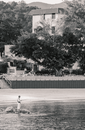 A person is standing on rocks by the edge of a body of water, holding a fishing rod. In the background, there is a sandy area, trees, and several buildings, including a multi-story structure with a tiled roof. The surrounding environment includes vegetation and a few outdoor settings with chairs and tables.