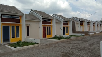 A row of small, modern single-story houses featuring bold, contrasting colors of yellow and brown around the windows and doors. The houses have uniform design with sloped roofs and are aligned neatly along a paved street. There are small patches of greenery in front of each house.