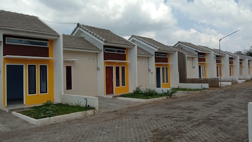 A row of small, modern single-story houses featuring bold, contrasting colors of yellow and brown around the windows and doors. The houses have uniform design with sloped roofs and are aligned neatly along a paved street. There are small patches of greenery in front of each house.