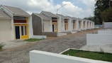 A row of identical small, single-story houses with tiled roofs and vibrant yellow door frames. The houses are aligned neatly along a paved road, with some greenery in the foreground and fenced off areas. The architecture appears modern and minimalistic, designed for functionality. The sky is partly cloudy, contributing to a neutral and calm setting.