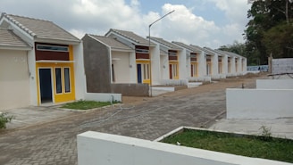 A row of identical small, single-story houses with tiled roofs and vibrant yellow door frames. The houses are aligned neatly along a paved road, with some greenery in the foreground and fenced off areas. The architecture appears modern and minimalistic, designed for functionality. The sky is partly cloudy, contributing to a neutral and calm setting.