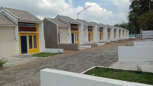 A row of identical small, single-story houses with tiled roofs and vibrant yellow door frames. The houses are aligned neatly along a paved road, with some greenery in the foreground and fenced off areas. The architecture appears modern and minimalistic, designed for functionality. The sky is partly cloudy, contributing to a neutral and calm setting.