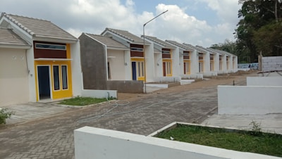 A row of identical small, single-story houses with tiled roofs and vibrant yellow door frames. The houses are aligned neatly along a paved road, with some greenery in the foreground and fenced off areas. The architecture appears modern and minimalistic, designed for functionality. The sky is partly cloudy, contributing to a neutral and calm setting.