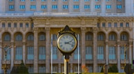 A large ornate clock, labeled 'Parliament,' is the focal point in front of a grand neoclassical building with a series of arched windows and intricate architectural details. The building's facade is composed of stone, featuring columns and a classical style. The scene is outdoors, with a clear sky and visible greenery surrounding the area.