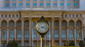 A large ornate clock, labeled 'Parliament,' is the focal point in front of a grand neoclassical building with a series of arched windows and intricate architectural details. The building's facade is composed of stone, featuring columns and a classical style. The scene is outdoors, with a clear sky and visible greenery surrounding the area.
