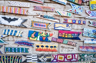 A collection of colorful, abstract fish-shaped wooden pieces is mounted on a textured stone wall. Each piece features unique patterns and vibrant colors, including stripes, dots, geometric shapes, and artistic symbols.