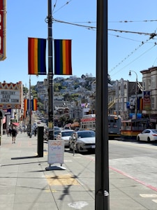 A vibrant street scene depicting a busy urban area with several rainbow flags adorning lamp posts, suggesting a sense of inclusivity or a celebration. The street is lined with cars and buildings, with a visible bus and pedestrians walking along the sidewalk. A signboard is visible in the foreground advertising a local business.