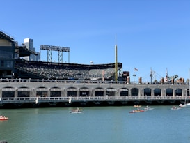 A baseball stadium with an outfield view is situated beside a body of water. Several people are kayaking on the water. The stadium is partially full, with flags flying above the stands. There is a large, yellow foul pole on the right side, and the skyline of a city is visible in the background.