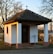 A small white chapel with a dark tiled roof and a cross on top, set against a backdrop of trees in a serene outdoor setting. The structure features two white columns and is situated near a quiet roadside.