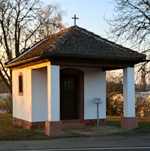 A small white chapel with a dark tiled roof and a cross on top, set against a backdrop of trees in a serene outdoor setting. The structure features two white columns and is situated near a quiet roadside.