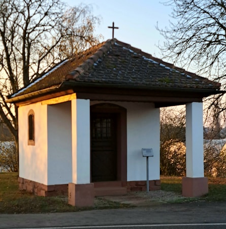 A small white chapel with a dark tiled roof and a cross on top, set against a backdrop of trees in a serene outdoor setting. The structure features two white columns and is situated near a quiet roadside.