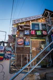 A quirky, eclectic building features a variety of signs and advertisements with vibrant colors and playful designs. Hanging bags with different brand logos adorn the facade, and the structure is decorated with lights and a small white cat figure in a window box. The scene includes visible wires, stairs, and a clear blue sky, imparting a whimsical, artistic vibe.