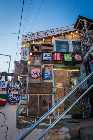 A quirky, eclectic building features a variety of signs and advertisements with vibrant colors and playful designs. Hanging bags with different brand logos adorn the facade, and the structure is decorated with lights and a small white cat figure in a window box. The scene includes visible wires, stairs, and a clear blue sky, imparting a whimsical, artistic vibe.