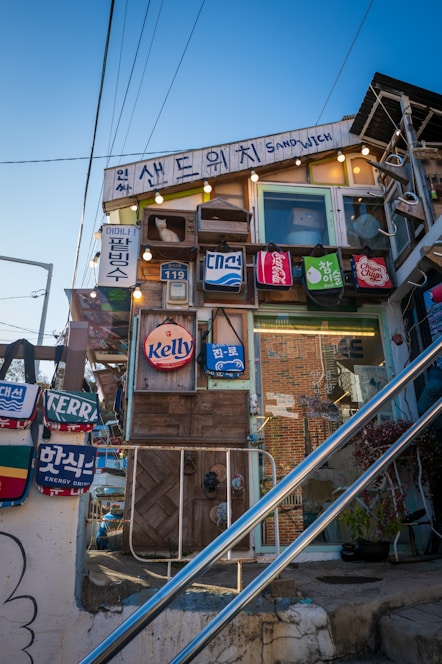 A quirky, eclectic building features a variety of signs and advertisements with vibrant colors and playful designs. Hanging bags with different brand logos adorn the facade, and the structure is decorated with lights and a small white cat figure in a window box. The scene includes visible wires, stairs, and a clear blue sky, imparting a whimsical, artistic vibe.