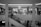 A black and white image of a shopping mall interior featuring a clothing store on the upper level with mannequins and clothing racks visible through the glass. Shoppers are walking on an escalator, moving both up and down. The mall is bright with circular ceiling lights, and a prominent sports brand logo is visible on the lower left.