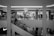 A black and white image of a shopping mall interior featuring a clothing store on the upper level with mannequins and clothing racks visible through the glass. Shoppers are walking on an escalator, moving both up and down. The mall is bright with circular ceiling lights, and a prominent sports brand logo is visible on the lower left.