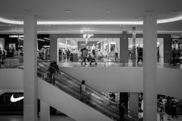 A black and white image of a shopping mall interior featuring a clothing store on the upper level with mannequins and clothing racks visible through the glass. Shoppers are walking on an escalator, moving both up and down. The mall is bright with circular ceiling lights, and a prominent sports brand logo is visible on the lower left.