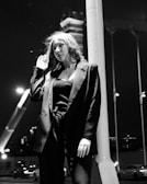 A black and white photograph of a person with long hair wearing a blazer and standing against a pole at night. The background shows blurred city lights and a tall structure, possibly a bridge or building.