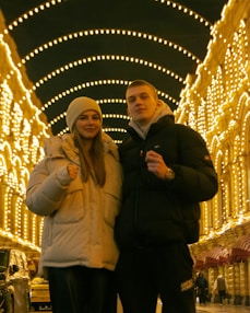A young couple stands under a canopy of glowing string lights in an outdoor setting. They are dressed warmly with puffer jackets, and the scene is illuminated with warm yellow and gold tones from the lights.