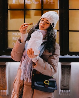 A woman in a winter coat and beanie is holding a paper-wrapped snack, likely eating on the go. She stands in front of a window displaying some text, with snowflakes lightly dusting her hat and shoulders.