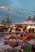 A festive outdoor amusement area with colorful decorations and lights. There's a small merry-go-round ride shaped like airplanes in the foreground. People dressed in winter clothing walk around the snow-covered ground. A tent-like structure and various stalls are visible, adorned with garlands, beneath the festive string lights.