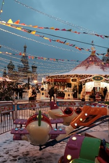 A festive outdoor amusement area with colorful decorations and lights. There's a small merry-go-round ride shaped like airplanes in the foreground. People dressed in winter clothing walk around the snow-covered ground. A tent-like structure and various stalls are visible, adorned with garlands, beneath the festive string lights.