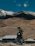 A rider on a motorcycle pauses on a mountain overlook with expansive Colorado peaks in the background under a clear blue sky.